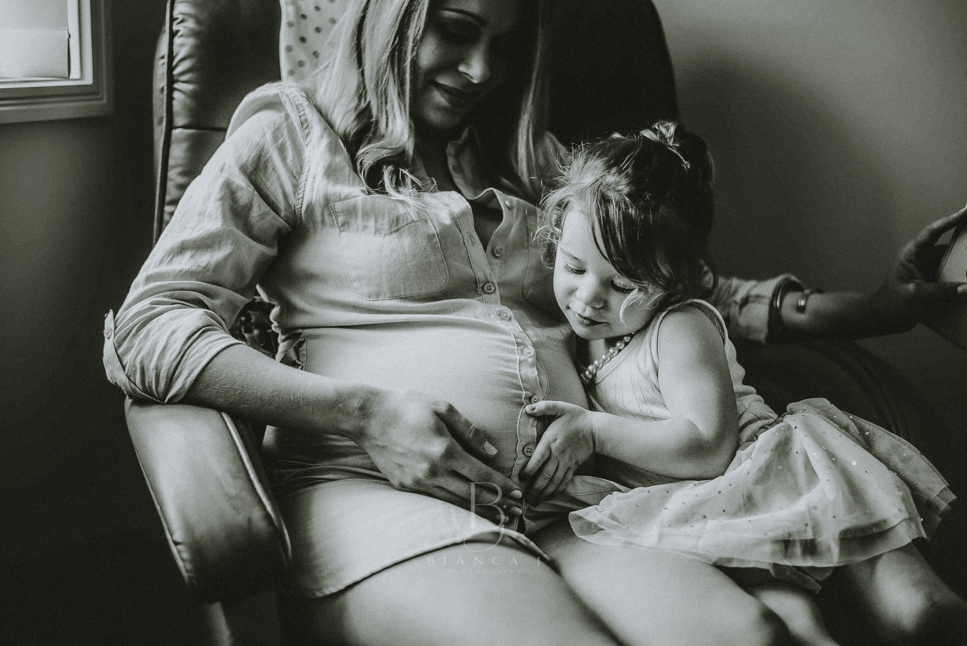 Toddler cuddling mum's bump in an armchair at home